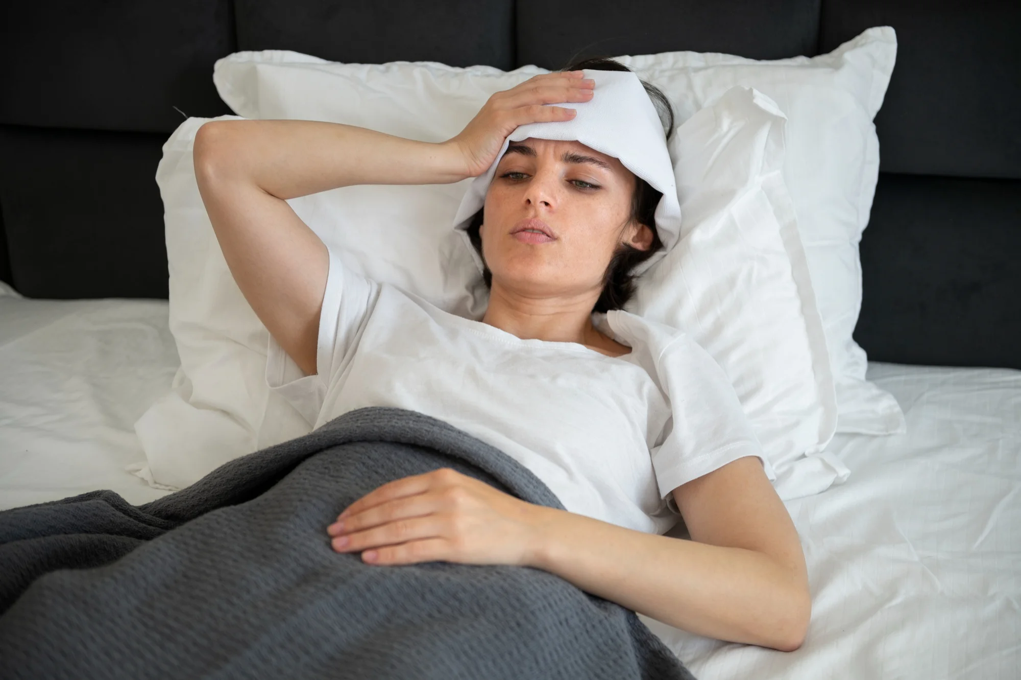 A woman lying in bed holds her head with a cold compress, appearing unwell, which may represent symptoms like headache, fever, or fatigue commonly linked to illness or poor sleep quality.