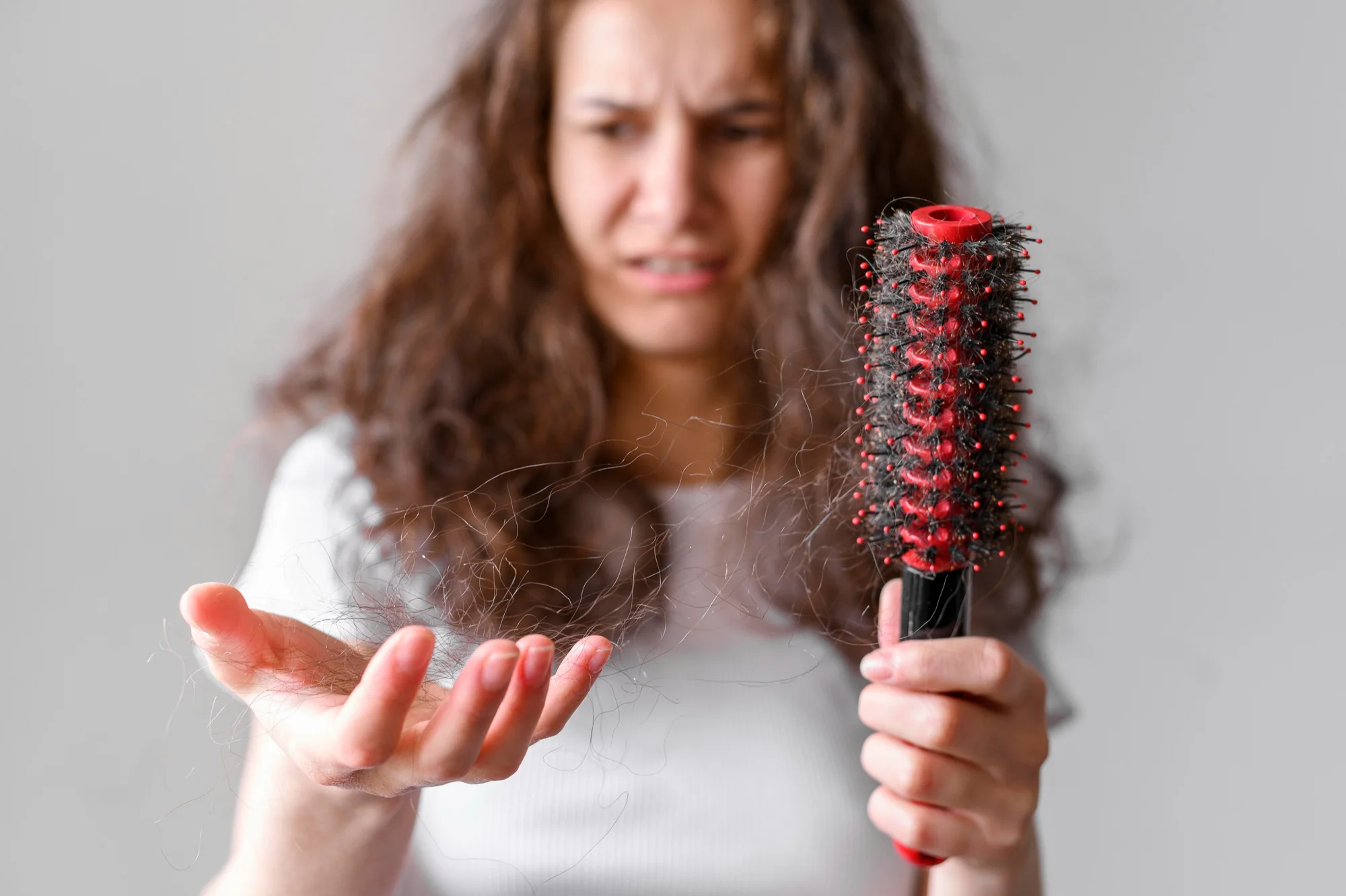 A woman holding a hairbrush with visible hair fall and frizzy strands in her hand, representing the comparison of Batana Oil vs Rosemary Oil for treating frizzy, damaged hair and improving hair strength and scalp health.