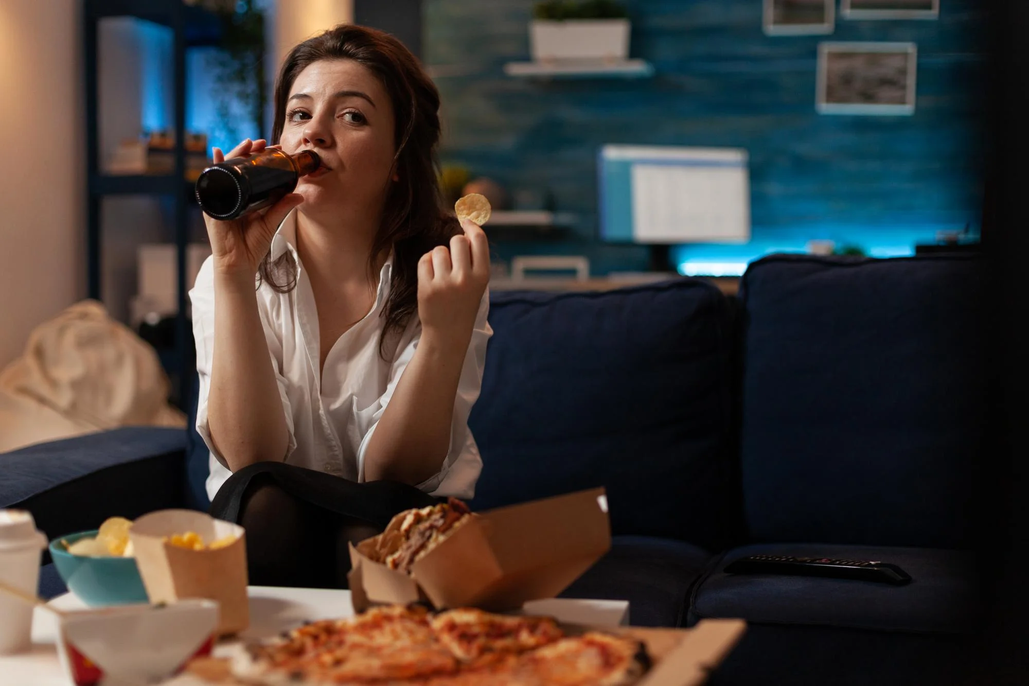 A woman sitting on a couch at night eating fast food and drinking a beverage, illustrating unhealthy eating habits and lifestyle factors that are commonly linked to sleep apnea and weight gain.