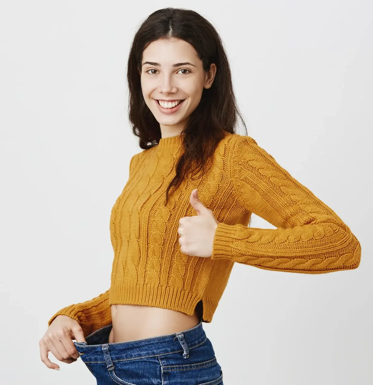 A smiling woman wearing loose jeans gives a thumbs-up gesture, symbolizing successful weight loss, improved health, and positive lifestyle changes.